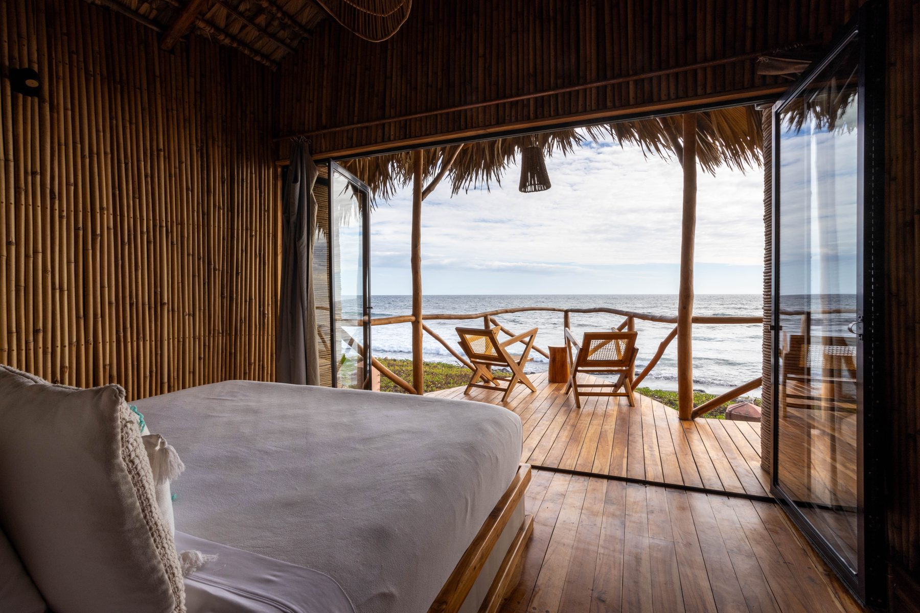 Inside a beachfront treehouse — king bed facing a deck with two chairs and the Pacific breaking just beyond the rail