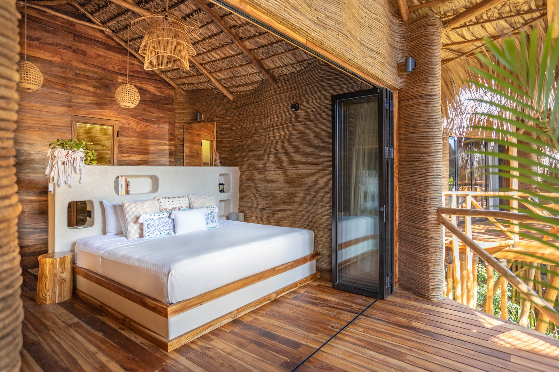Inside a jungle treehouse — king bed against a sculpted headboard, rattan pendants, folding doors open to a deck in the palms