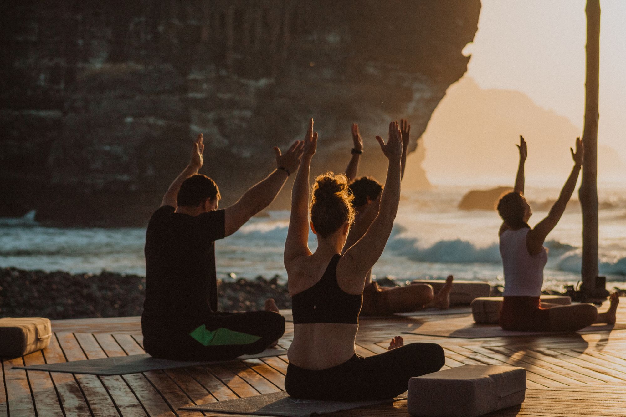 Guests on mats at sunset, arms raised, on the open deck above the surf